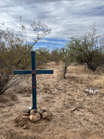 A homemade cross stands in the ground in a desert area surrounded by rocks and cacti. The location is in Southern Arizona near the U.S. - Mexico border.